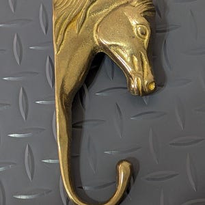 May include: A polished brass wall hook shaped like a horse head and neck. The horse is depicted in profile, facing right, with detailed mane textures along the top edge. The lower portion of the piece curves into a functional hook for hanging items.