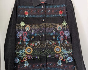 Op de afbeelding: Een donker antracietgrijze jas met knopen en een levendig, veelkleurig bloemen- en geometrisch borduurpatroon aan de voorzijde. De jas heeft een klassieke kraag, lange mouwen en een rij donkere knopen in het midden.