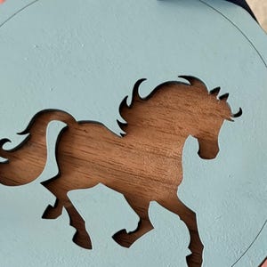 May include: A circular light blue wooden ornament featuring a cutout silhouette of a horse in the center. The natural brown wood grain is visible through the cutout. A dark blue ribbon is attached to the top for hanging purposes.