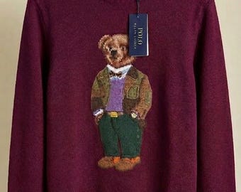 May include: A deep burgundy long-sleeve crewneck sweater featuring a central graphic of a teddy bear wearing a plaid blazer, purple sweater, and green trousers. The sweater has a ribbed collar, cuffs, and hem, with the letters RL embroidered in gold.