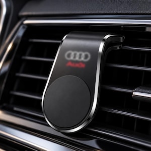 May include: A black magnetic car phone mount with a brushed metal finish and silver edges, featuring the Audi logo in white and red. The mount is attached to a horizontal vehicle air vent, showcasing a circular magnetic surface for device holding.