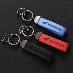 May include: Three keychains featuring a metal ring and a rectangular strap in blue, red, and black. Each strap displays the Honda logo and text. The metal hardware is dark grey, and the straps have visible stitching along the edges.