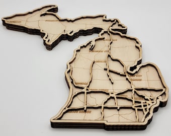 Peut inclure: Une carte en bois superposée de l'État du Michigan, avec des noms de villes gravés tels que Marquette, Traverse City, Cadillac et Benton Harbor. La construction en bois clair montre des frontières d'État et des divisions régionales.