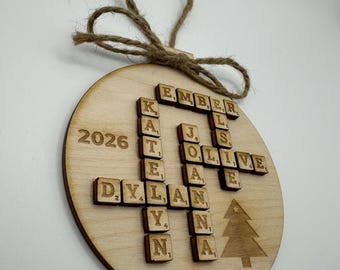Peut inclure: Une décoration ronde en bois avec des lettres en relief disposées en mots croisés formant les prénoms Ember, Katelyn, Dylan, Jo et Olive. L'année 2026 est imprimée à gauche, un sapin à droite, avec une boucle en ficelle de jute.