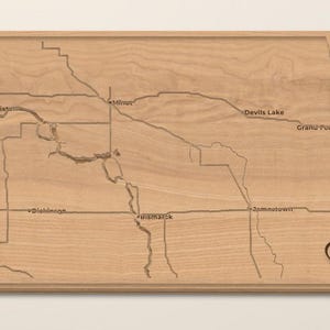 May include: A light-toned wooden wall art piece shaped like the state of North Dakota, featuring laser-engraved state borders, major rivers, and city names including Williston, Minot, Devils Lake, Grand Forks, Dickinson, Bismarck, Jamestown, and Fargo.