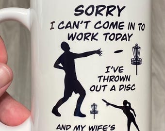 Peut inclure: Un mug en céramique blanc avec le texte noir : SORRY I CAN'T COME IN TO WORK TODAY I'VE THROWN OUT A DISC AND MY WIFE'S TEE'D OFF. Le motif présente des silhouettes noires de deux personnes lançant des disques vers des paniers de disc golf.