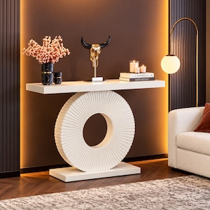 May include: A white console table with a rectangular top and a large, circular, ribbed pedestal base. On the table sits a black marble vase with pink flowers, a gold bull skull sculpture, and two books titled Kinfolk and Louis Vuitton.