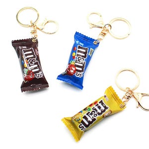 May include: Three keychains featuring miniature replicas of M&M's candy wrappers in brown, blue, and yellow. Each wrapper has the M&M's logo and a character graphic printed on the front. The keychains are attached to gold-toned metal rings and clasps.