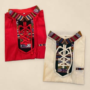Peut inclure: Deux chemises, une rouge et une blanc cassé, présentant chacune un panneau de poitrine noir avec un laçage. Les cols et les panneaux de poitrine sont ornés de bandes de tissu tissé aux motifs colorés. Chaque chemise possède une petite poche.