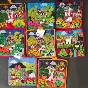 Könnte beinhalten: Eine Sammlung von acht quadratischen Topflappen aus Stoff mit farbenfrohen gestickten Landschaften, die Lamas, Berge, Bäume und Häuser zeigen. Jedes Stück hat einen gehäkelten Rand und eine Aufhängeschlaufe. Ein weißes Studio Artesanos-Etikett ist an einem Topflappen befestigt.