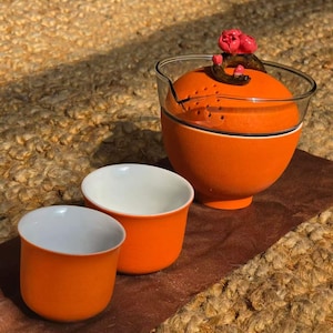 May include: A bright orange tea set consisting of a glass-topped teapot with a dark brown branch handle and pink flower accents, accompanied by two matching orange tea cups with white interiors, all featuring a smooth, matte finish.