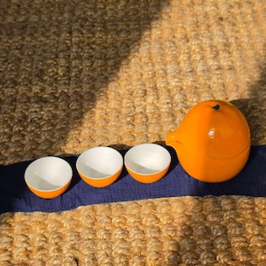 May include: A ceramic tea set consisting of three small, round bowls with white interiors and bright orange exteriors, accompanied by a matching orange teapot shaped like a whole orange, all arranged on a dark blue fabric runner.