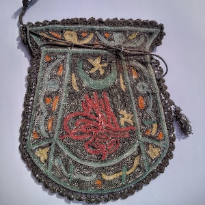 May include: A dark brown drawstring pouch featuring intricate needlework embroidery. The design includes a central red calligraphic emblem, a green crescent moon, and a yellow star, accented with orange and light blue floral-inspired motifs.
