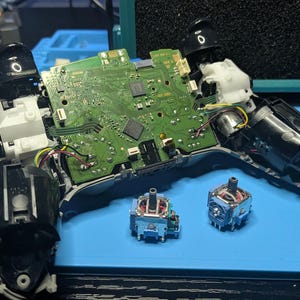 May include: A disassembled video game controller with its green circuit board exposed, featuring two detached analog joystick modules placed in front of the main housing, revealing internal black plastic components and wiring.
