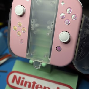 以下が含まれることがあります： パステルピンクのゲームコントローラーグリップ。白いジョイスティックとボタン、半透明の中央セクションを備えています。コントローラーは、赤いNintendoロゴと白い「MINTY'S CUSTOMS」の文字が表示されたライトグリーンのスタンドに設置されています。