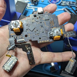 May include: A black printed circuit board assembly featuring two yellow analog joystick modules, a central processor, and two smaller connected circuit boards with metallic components, all linked by thin gray ribbon cables and wires.