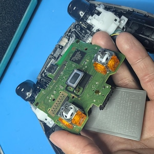 以下が含まれることがあります： 分解されたビデオゲームコントローラー。緑色の回路基板、2つのオレンジ色のスティックモジュール、中央の黒い長方形の部品、銀色のバッテリーパックが、青い作業台の上で修理作業中に露出している様子。