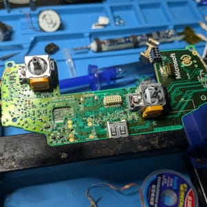 Puede incluir: Una placa de circuito impreso verde de un mando de juego Microsoft, fijada en un tornillo de banco de metal azul. La placa cuenta con dos módulos de joystick analógicos con bases amarillas, un puerto USB plateado y varios componentes.