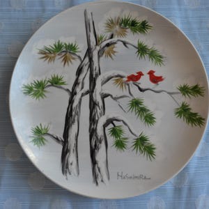 May include: A white ceramic plate with a hand-painted design of two red birds perched on a pine tree branch. The plate has a black and white illustration of the tree with a snowy background. The artist's signature, "Hashimura", is visible in the lower right corner.