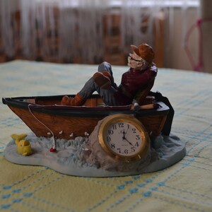 May include: A decorative figurine featuring a person wearing a brown cap and red shirt sitting in a wooden rowboat. A round analog clock with a gold frame is embedded in the base, which also includes a small yellow fish and a fishing line.