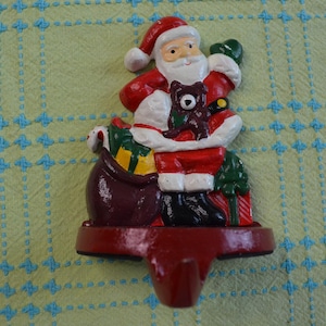 May include: A cast iron stocking holder featuring a painted Santa Claus figure in a red suit, holding a brown teddy bear. Santa stands next to a dark red sack filled with yellow and green gifts, with a small green tree at the base and a hook.