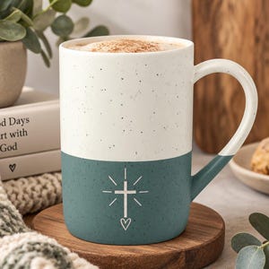 May include: A two-toned ceramic mug with a speckled cream upper half and a solid teal lower half. The teal section features a white graphic of a cross with radiating lines and a small heart at the base. The mug has a curved handle on the right side.