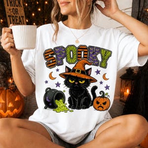 Könnte beinhalten: Ein weißes T-Shirt mit einer Grafik eines schwarzen Katers mit orangefarbenem Hexenhut, einem schwarzen Kessel mit auslaufender grüner Flüssigkeit und einem kleinen orangefarbenen Kürbis. Das Wort SPOOKY steht darüber in orange, grün und lila.