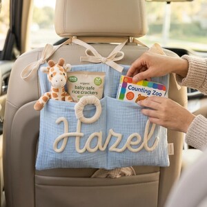 May include: A light blue fabric car seat organiser featuring the name Hazel in white rope lettering. The organiser has two pockets containing a plush giraffe toy, a package of organic baby-safe rice crackers, and a book titled Counting Zoo.