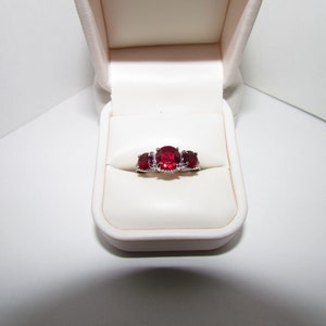 May include: A silver-toned ring featuring three round, deep red faceted stones set in a row. The center stone is larger than the two flanking stones. The ring is displayed inside a cream-colored, hinged jewelry box with a soft, padded interior.