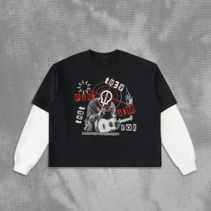 May include: A black long-sleeve shirt with white undersleeves featuring a graphic print of a person playing an acoustic guitar. The design includes a red target symbol, the text "PART THAT THEY AIM FOR," and a string of letters at the bottom.