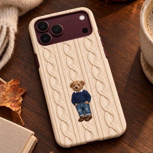 May include: A cream-colored phone case with a textured cable knit pattern. The case features a central graphic of a brown teddy bear wearing a navy blue sweater, a white collared shirt, blue denim jeans, and brown shoes, set against a dark camera cutout.