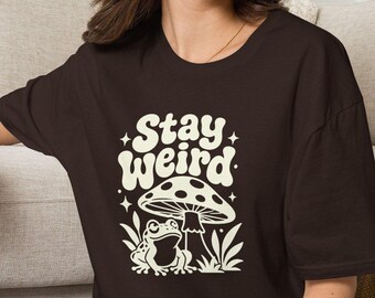 May include: A dark brown short-sleeve t-shirt featuring a cream-colored graphic print of a frog sitting under a mushroom with the text Stay Weird, paired with blue denim jeans and white sneakers, creating a casual and comfortable outfit.