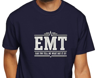 May include: A navy blue short-sleeve t-shirt featuring a white graphic design on the chest. The design includes the letters EMT in a bold font, with the text CAN YOU TELL ME WHAT DAY IT IS? underneath, framed by horizontal lines and a pulse icon.