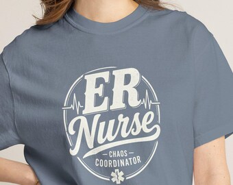 May include: A slate blue short-sleeved t-shirt featuring a white circular graphic on the chest. The text reads ER Nurse, Chaos Coordinator, with a heartbeat line and a small star of life symbol centered within the circular design.