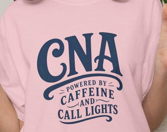 May include: A light pink short-sleeved t-shirt featuring dark blue text that reads CNA POWERED BY CAFFEINE AND CALL LIGHTS. The shirt has a crew neckline and a relaxed fit, with the graphic centered on the chest in a bold, stylized font.
