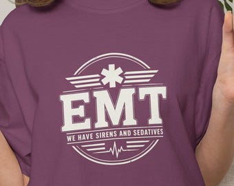 May include: A plum-colored short-sleeve t-shirt featuring a white circular graphic with the text "EMT" and "WE HAVE SIRENS AND SEDATIVES" centered on the chest. The design includes a star of life symbol and a heartbeat line within the circle.