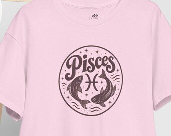 May include: A light pink short-sleeve t-shirt featuring a circular graphic on the chest. The graphic contains the word Pisces in a stylized font, the Pisces zodiac symbol, and two fish illustrations surrounded by small stars and wave lines.