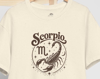 May include: A cream-colored short-sleeve t-shirt with a speckled texture featuring a dark brown graphic of a scorpion and the text Scorpio above the astrological symbol for the sign. A plain white baseball cap is draped over the wooden chair.