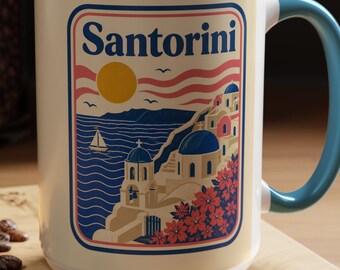 May include: A white ceramic mug with a light blue interior and handle, featuring a graphic illustration of Santorini with the text Santorini, a yellow sun, blue domed buildings, a sailboat on the sea, and red flowers within a rectangular frame.