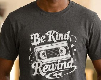 May include: A dark heather gray short-sleeve t-shirt featuring a white graphic print of a vintage VHS cassette tape in the center. The text "Be Kind" is printed above the cassette, and "Rewind" is printed below, accompanied by small stars.