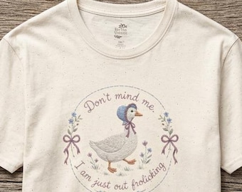 May include: A cream-colored short-sleeve t-shirt featuring a central graphic of a white goose wearing a blue bonnet and a pink bow. The illustration is encircled by a floral wreath and the text, "Don't mind me. I am just out frolicking."