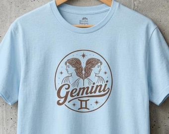 May include: A light blue short-sleeve t-shirt featuring a circular brown graphic on the chest. The design depicts two profile faces representing the Gemini zodiac sign, the word Gemini in a cursive script, and the Roman numeral II symbol.