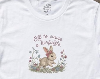May include: A white short-sleeve t-shirt featuring a graphic of a brown rabbit with a pink bow in a field of flowers. The text above the rabbit reads "Off to cause a kerfuffle." The shirt has a crew neckline and a Bertha Murphy brand label inside.