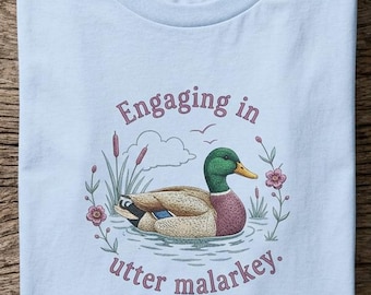 May include: A light blue t-shirt featuring a graphic of a mallard duck swimming in water, surrounded by cattails and small flowers. The text "Engaging in utter malarkey" is printed in a curved purple font above and below the duck illustration.