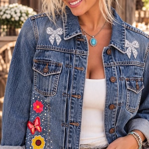May include: A medium-wash denim jacket featuring two silver rhinestone bow appliqués on the chest, a pink flower patch, a red butterfly patch, and a yellow sunflower patch on the right side, accented with scattered clear rhinestones.