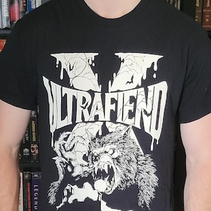 以下が含まれることがあります： 黒の半袖Tシャツで、大きな白いグラフィックプリントが施されています。デザインには、滴るようなひび割れたフォントで「ULTRAFIEND」という文字が描かれ、その下に鋭い爪と牙を持つ唸る狼男の詳細なイラストが配置されています。