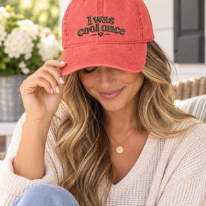 May include: A coral-colored baseball cap with a distressed finish featuring the embroidered dark green text "I was cool once" centered on the front panel, accompanied by a small heart icon and two horizontal lines beneath the lettering.