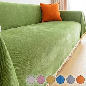 May include: A sage green sofa cover with a textured herringbone pattern and a decorative fringe along the bottom edge. A solid orange square throw pillow rests on the seat. Six small circular color swatches are displayed at the bottom.