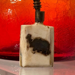 May include: A small, white, rectangular bottle with a metal top and a silver elephant design on the front. The bottle is sitting on a clear surface.