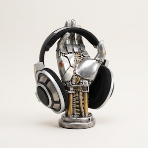 May include: A silver-colored robotic hand sculpture with visible mechanical joints, gold-toned wiring, and a circular base, functioning as a stand for a pair of black and silver over-ear headphones that are resting on the fingers of the hand.